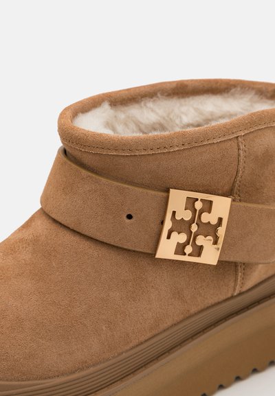 Smeđi antilop čizme do gležnja s mekšanim unutarnjim dijelom, s širokom trakom i zlatnim dekorativnim detaljem u obliku kopče. Debela, teksturirana potplate.