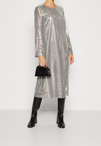 Robe à manches longues en sequins argentés, coupe loose, col rond, associée à un petit sac à main noir en texture crocodile et des bottes noires jusqu'aux genoux.