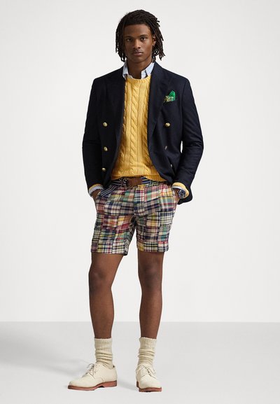 Blazer marine, pull en maille torsadée jaune, short à carreaux de différentes couleurs, chaussures beige et chaussettes crème. Un mouchoir de poche ajoute du détail.
