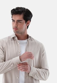 Camicia beige con bottoni, realizzata in un tessuto leggermente testurizzato e con maniche lunghe, indossata sopra una T-shirt bianca. Presenta un colletto classico e polsini abbottonati.