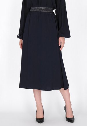 La modelo lleva un vestido azul marino oscuro, hasta media pantorrilla, de manga larga y con una cintura texturizada, combinado con zapatos negros de tacón.