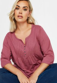 Vaaleanpunainen pitkähihaisen yläosan, jossa on henley-kaulus, poimutettu etuosa ja napit. Pehmeä kangas ja rento istuvuus.