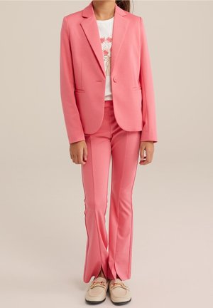 Completo rosa con blazer su misura e pantaloni svasati, realizzato in tessuto liscio. I pantaloni presentano strisce laterali. Camicia bianca sotto.