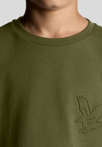 Oliven grøn bomulds t-shirt med rund hals og et præget ørnelogo på venstre brystområde. Blødt, struktureret stof.