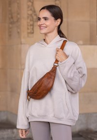 Felpa oversized grigia chiara con tasca frontale, abbinata a pantaloni aderenti chiari. Borsa a tracolla in pelle marrone con dettaglio zip.