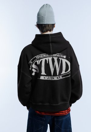 Zwarte hoodie met een grote witte "STWD" opdruk op de achterkant, voorzien van contrasterende stiksels. Draagt daarbij een lichtblauwe gebreide muts.