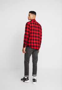 Camisa de franela a cuadros rojos y negros con mangas largas, puños con botones y corte recto, combinada con jeans gris oscuro y zapatillas deportivas.