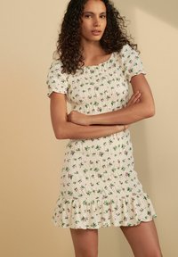 Robe florale blanche avec des feuilles vertes et des fleurs roses. Texture smockée et manches courtes, avec un ourlet à volants.