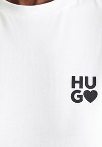 Valge puuvillane T-särk, millel on must trükitud graafika, millel on tekst "HUGO" ja südame sümbol, ümar kaelus ja lühikesed varrukad.