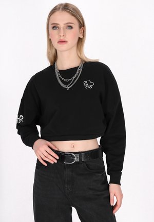 Zwarte cropped sweatshirts met geborduurd hart en pootontwerp; gecombineerd met hoog uitgesneden zwarte broek en een glanzende zwarte riem.