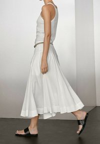 Jupe plissée blanche avec un ourlet large et une taille ajustée, associée à un débardeur gris et des sandales à glisser noires. Texture de tissu lisse.