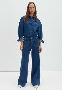 Femme debout portant un jean denim bleu foncé à jambes larges et une chemise en denim à manches longues assortie avec les manches retroussées, devant un fond clair uni.