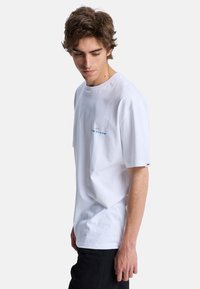 Jeune homme aux cheveux bruns portant un t-shirt blanc Quiksilver et un pantalon noir, regardant vers le bas sur un fond uni.