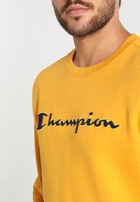Felpa gialla in cotone, con un grande logo "Champion" ricamato in nero e rosso. Collo a giro e polsini a coste.
