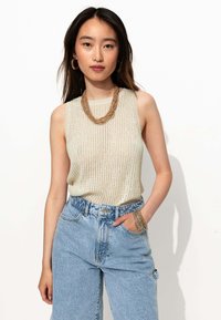 Haut sans manches côtelé beige avec un col rond, associé à un jean en denim bleu clair taille haute. Les accessoires comprennent un collier en or superposé et des bracelets.