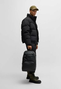 Black padded jacket, black backpack with a textured finish, olive cap, and green boots. The backpack has front zip pockets and a logo.