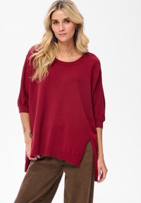 Czerwony sweter wykonany na drutach z okrągłym dekoltem, rękawami trzyczworęcznymi i bocznymi rozcięciami; noszony na brązowych spodniach. Charakteryzuje się miękką fakturą i luźnym krojem.