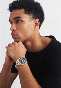 Gold watch with blue face, metallic band, round dial, luminous hands, and chronograph features. Model wearing a black shirt, hands clasped.