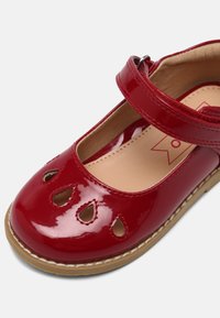 Scarpa in pelle verniciata rossa con punta rotonda, cinturino regolabile e modelli a goccia ritagliati sulla parte anteriore, dotata di una fodera interna beige.