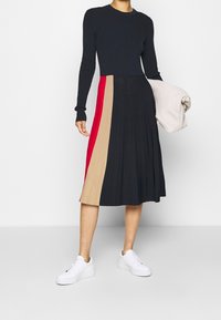 Haut en maille noir à manches longues, associé à une jupe plissée en blocs de couleurs comprenant des sections noir, beige et rouge, porté avec des baskets blanches.