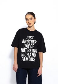 Czarna, oversize koszulka z krótkim rękawem z odważnym białym napisem "KOLEJNY DZIEŃ NIE BYCIA BOGATYM I FAMOUS." Luźny krój.