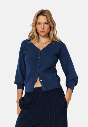 Cardigan bleu marine avec un décolleté en V, des manches trois-quarts et des boutons dorés. Texture côtelée avec une coupe décontractée, associé à un pantalon à rayures.