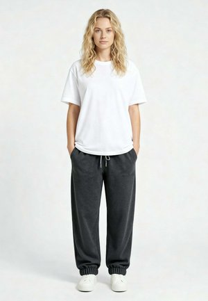 Femme aux cheveux blonds bouclés portant un t-shirt blanc, un pantalon de survêtement gris foncé avec cordon, et des baskets blanches, debout les mains dans les poches.