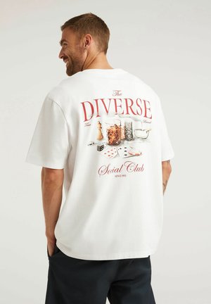 Mężczyzna w białej koszulce z nadrukiem o tematyce hazardowej, zawierającym karty, żetony, kości i napój, z napisem "The Diverse Social Club Since 1993".