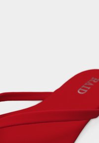 Κόκκινο σανδάλι με ψηλό τακούνι και λεπτό λουράκι, με ορατό ασημί όνομα μάρκας στην εσωτερική σόλα, σε απλό φωτεινό φόντο.