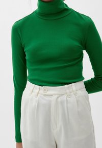 Grüner langärmeliger Rollkragenpullover aus glattem Stoff, kombiniert mit hoch geschnittenen cremfarbenen Hosen mit seitlichen Falten und einem Knopfverschluss.