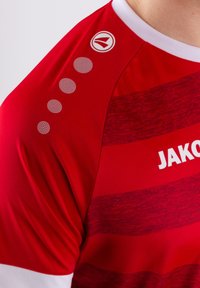 Rotes Sporthemd mit gestreifter Textur, ausgestattet mit weißen Akzenten und einem Logo auf der Schulter. Rundmuster-Detail in der Nähe des Kragens.