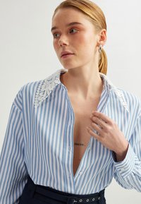 Camicia a righe blu e bianche con colletto in pizzo, maniche lunghe, chiusura con bottoni e scollatura profonda a V. Il tessuto appare liscio e leggero.
