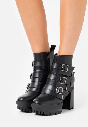 Stivaletti neri in pelle con tacchi robusti, suole spesse, cerniera laterale e tre cinturini regolabili con fibbia, indossati su gambe nude.