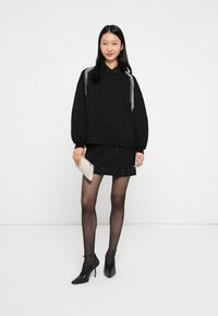 Žena v čiernom oversize svetríku s perlovými detailmi, čiernej minisukni, priehľadných čiernych pančuchách a čiernych lodičkách, ktorá drží biely clutch.