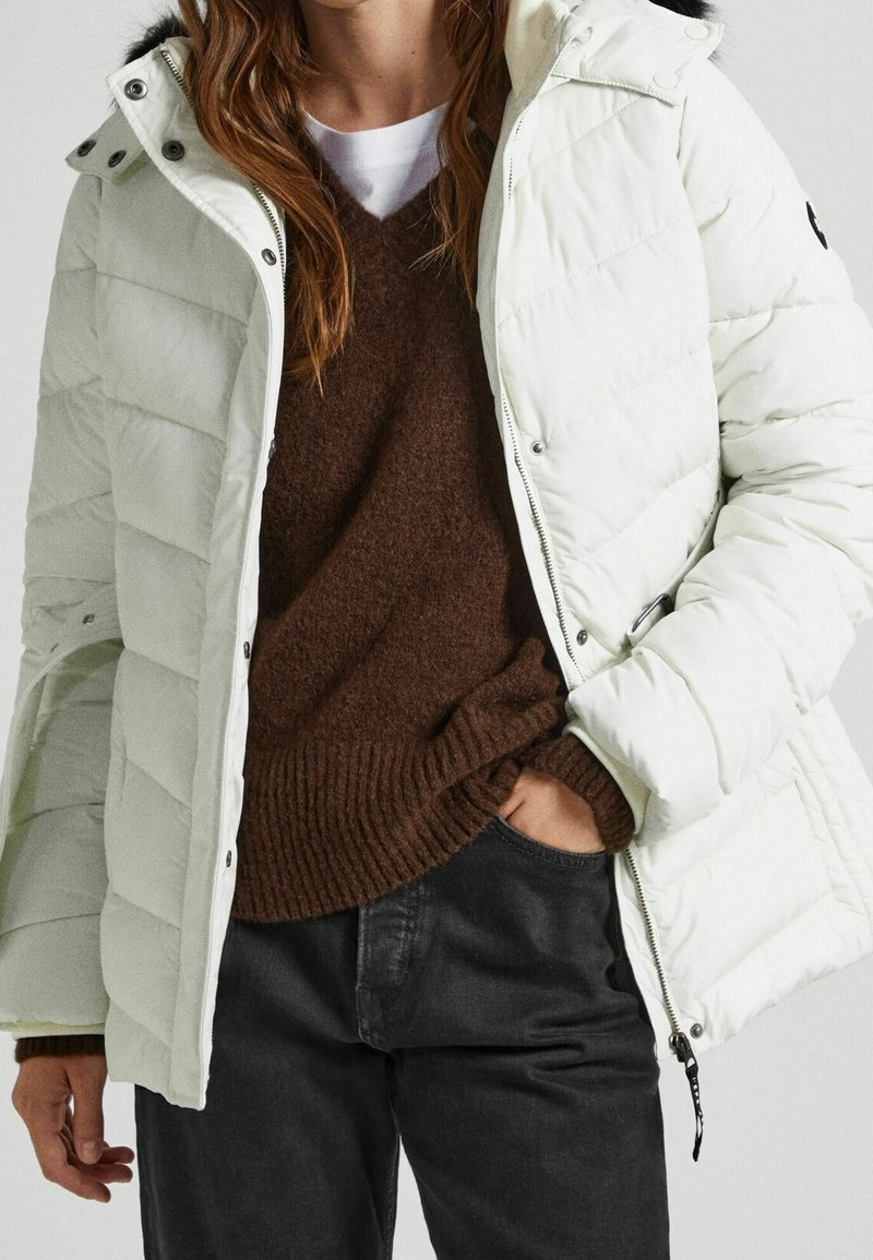 Veste d'hiver blanche matelassée avec capuche, équipée d'une fermeture éclair et de boutons-pression, portée sur un pull en tricot marron et un pantalon noir.
