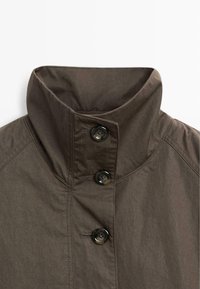 Brązowa kurtka z wysokim kołnierzem, z dwoma czarnymi guzikami i ukośnymi detalami przeszyć na tkaninie, o miękkiej teksturze i wyraźnej strukturze.