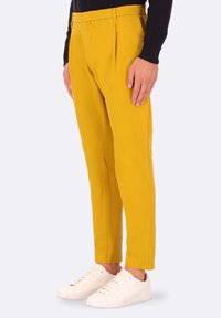 Pantaloni di cotone gialli con vestibilità su misura, caratterizzati da pieghe frontali e tasche laterali, abbinati a sneaker bianche.