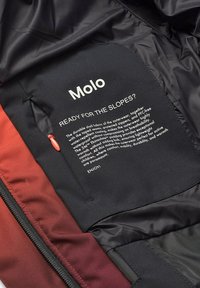 Tessuto interno nero con accenti rossi presenta un'etichetta di cura che indica "Molo" e istruzioni per la cura. Tasca con zip, materiale testurizzato.