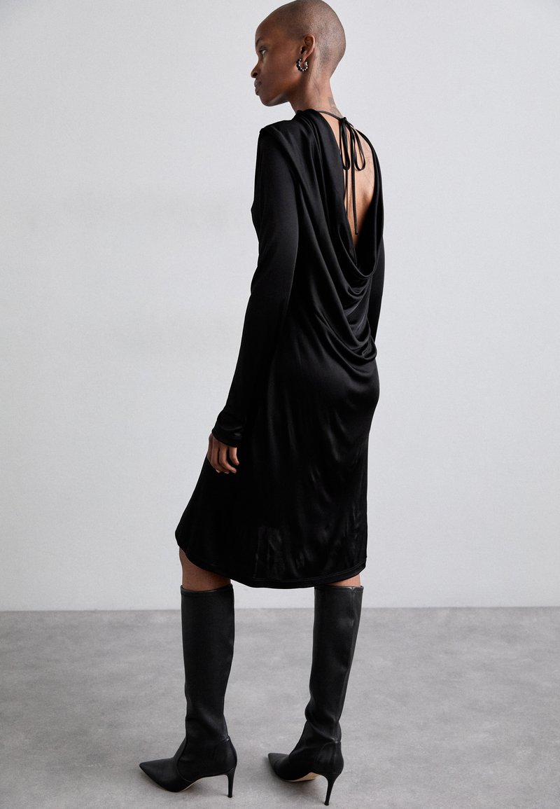 Black draped dress with long sleeves and an open back, made of smooth fabric. Paired with knee-high black pointed boots. Simple design.