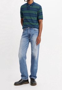 Man i blå- och grönrandig kortärmad polo, ljusblå raka jeans och svarta snörskor står mot vit bakgrund.