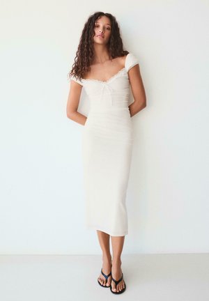 Mladá žena s kučeravými vlasmi v priliehavých bielych midi šatách s odhalenými ramenami a čiernych žabkách, stojaca pred jednoliatou bielou stenou.