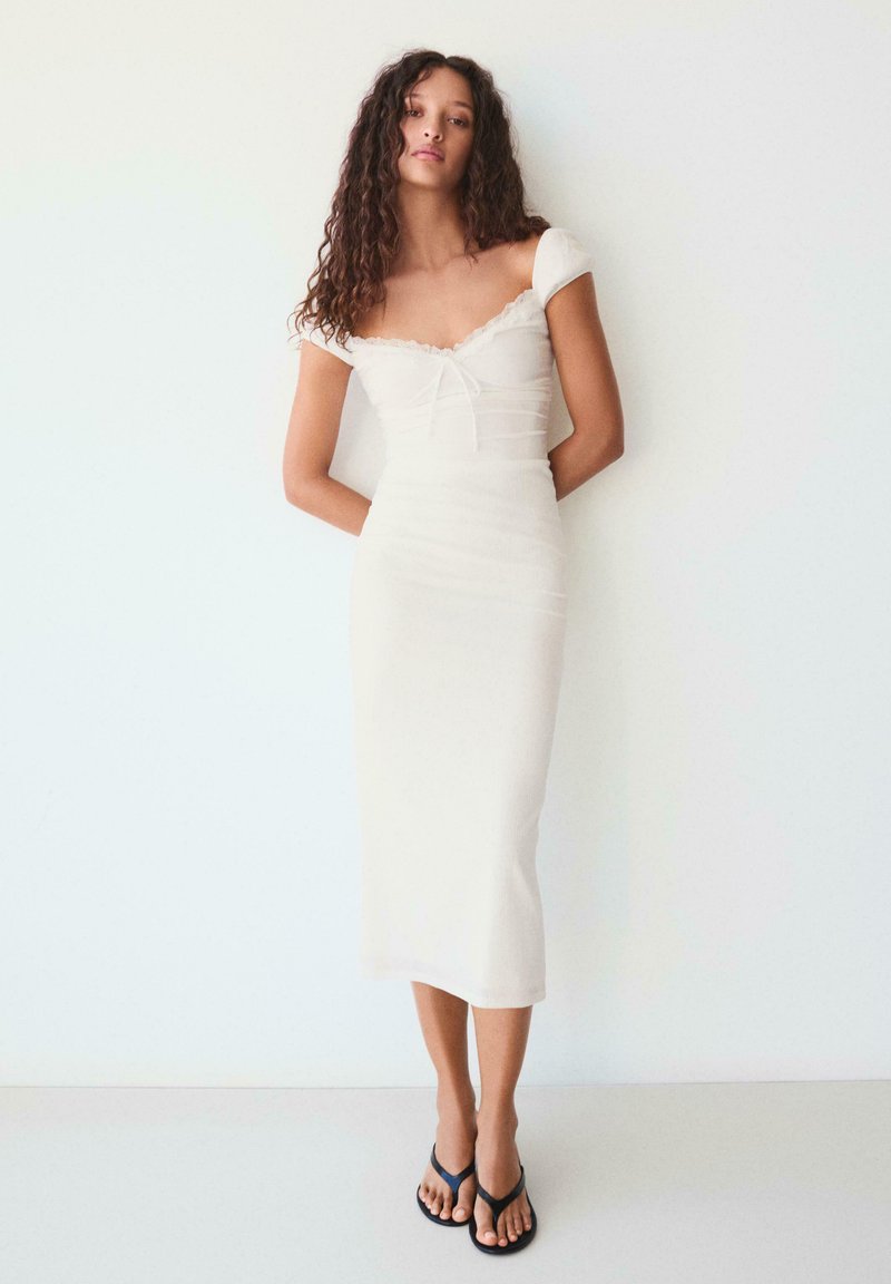 Fiatal nő göndör hajjal, fehér, vállakat szabadon hagyó, testhez simuló midi ruhát és fekete papucs szandált visel, egy sima fehér fal előtt áll.