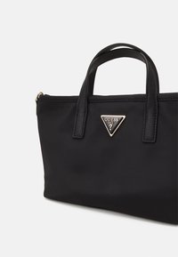 Czarna sztuczna torba tote z zaokrąglonymi uchwytami, wyposażona w trójkątny emblemat logo w kolorze złotym oraz gładką teksturę. Kompaktowy design zapewniający wszechstronność.