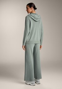 Set iz svetlo zelene rebraste pletenine, ki vključuje kapuco in široke hlače, sproščenega kroja, dopolnjen z belimi in sivimi tekaškimi čevlji.