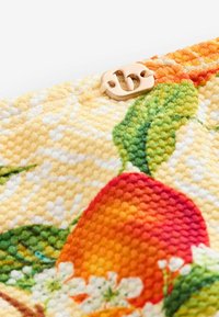 Nærbillede af tekstureret stof med farverigt frugt- og bladmotiv samt en lille beige mærkat med bogstaverne "b" og "s".