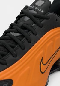 Sapatilha Nike Shox preta com um painel de amortecimento laranja, parte superior texturada e atacadores pretos. O logo da Nike é destacado em laranja.