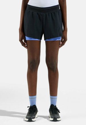 Sportowiec ubrany w czarne, warstwowe spodenki do biegania na niebieskich kompresyjnych spodenkach, jasnoniebieskie skarpety i czarne buty do biegania stojący na białym tle.