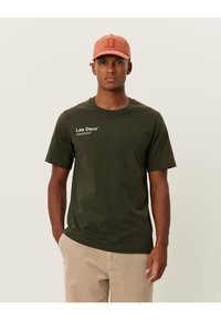 Olive green cotton t-shirt featuring short sleeves and a crew neck, with white text logo on the left chest, paired with beige corduroy pants.