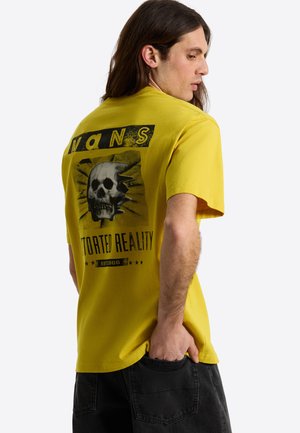 Mand med langt hår iført en gul Vans T-shirt med en kraniegrafik og teksten "Distorted Reality", hånden i lommen, vendt sidelæns.