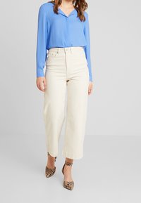 Modrá blůza s dlouhým rukávem a límečkem, doplněná o kalhoty s vysokým pasem v off-white barvě a širokými nohavicemi, a na nohou hadí vzorované sandály na kotníkovém popruhu.